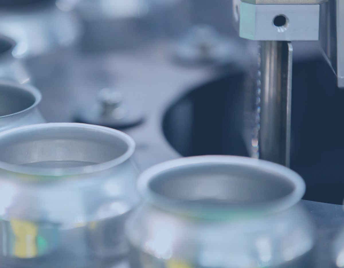 Close-up of an automated manufacturing line with metal containers positioned beneath a precision filling or processing machine in an industrial production setting.