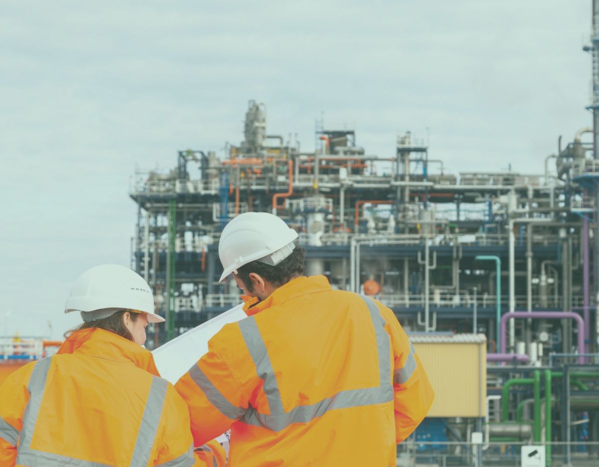 Two workers in high-visibility jackets and hard hats review plans at an industrial processing facility, with complex piping, platforms, and equipment structures in the background.