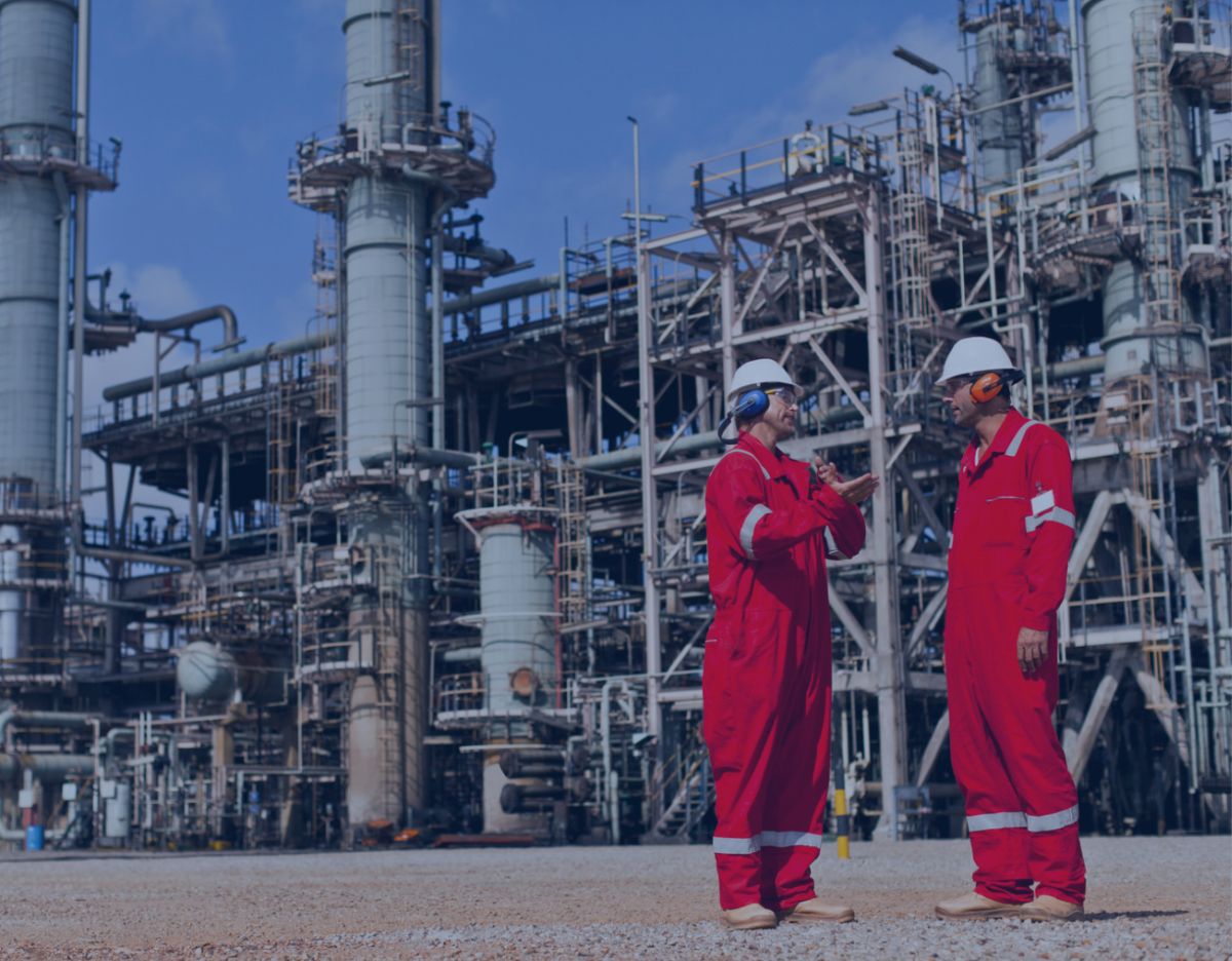 Two workers in red protective coveralls and hard hats stand at an industrial refinery site, discussing operations while one reviews information on a handheld device amid large processing units and piping.
