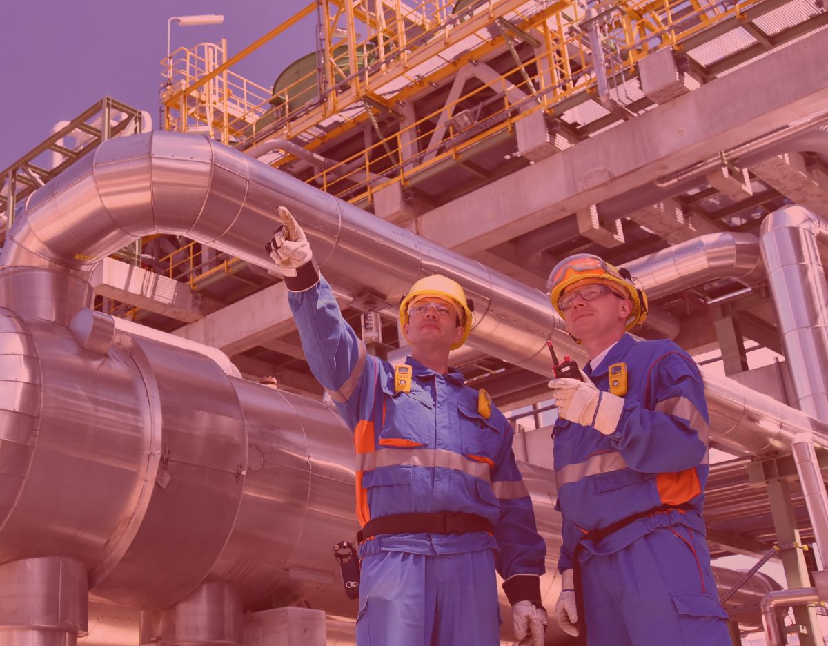 Two industrial workers in protective gear stand in a processing facility, reviewing equipment and communicating while one points toward large stainless steel piping and elevated structures.