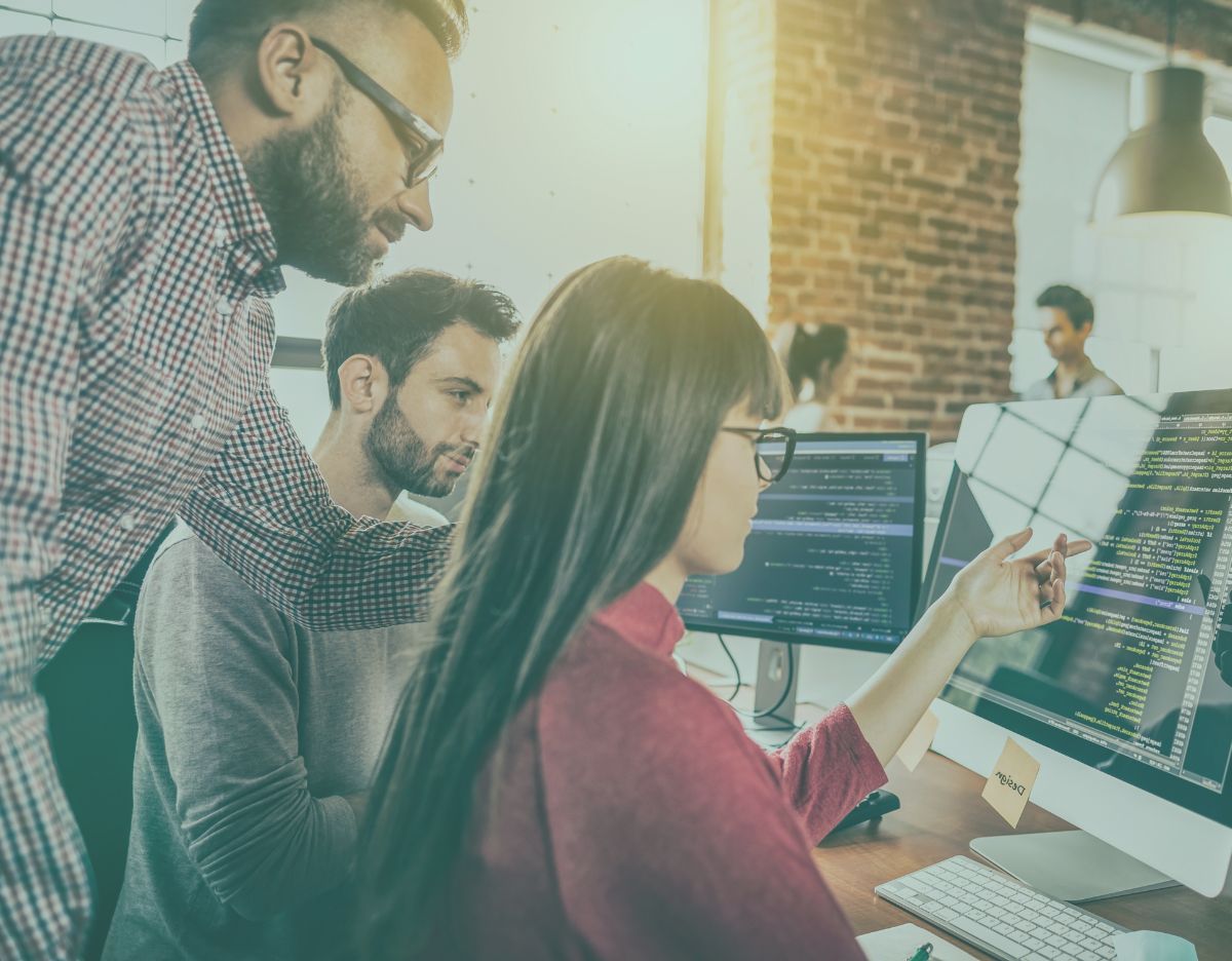 Team of professionals collaborating in an office, reviewing code and data on large desktop monitors during a technical discussion.