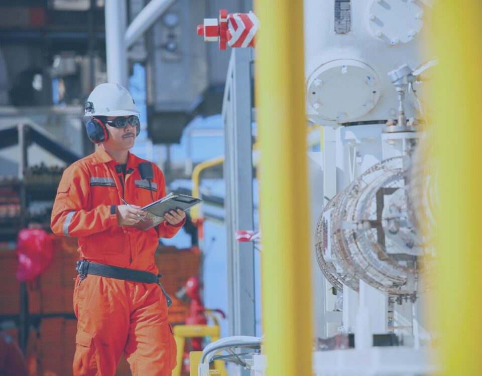 Worker in orange protective coveralls and a hard hat uses a tablet to inspect industrial piping and equipment inside a processing facility.