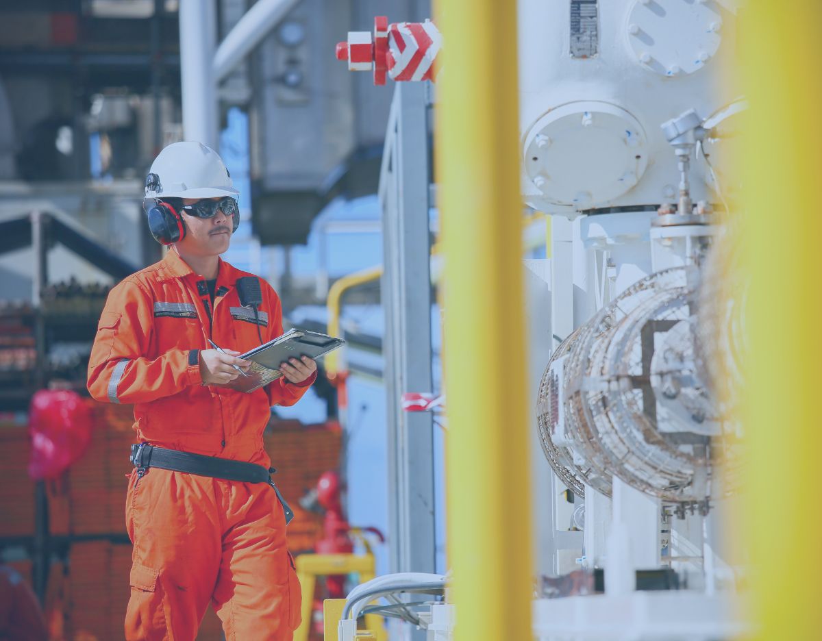 Worker in orange protective coveralls and a hard hat uses a tablet to inspect industrial piping and equipment inside a processing facility.