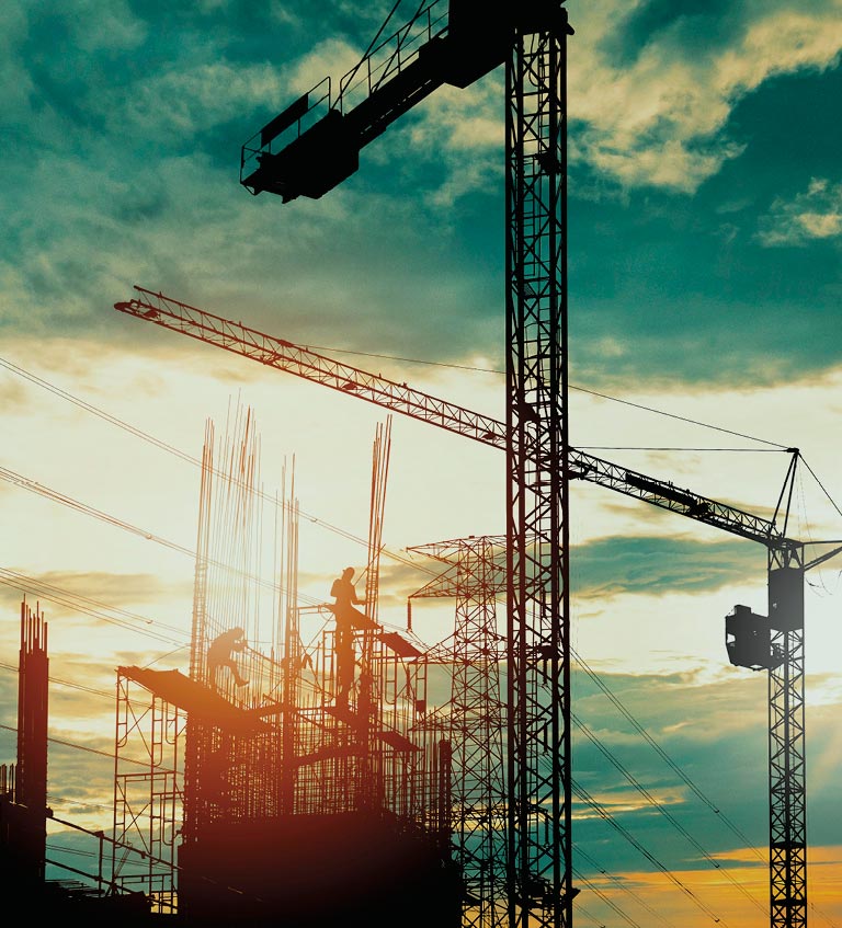 Silhouettes of construction cranes and workers atop a building framework at sunset under a partly cloudy sky.