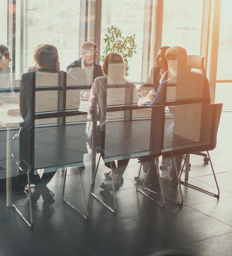 A group of people sit around a conference table in a glass-walled office, engaged in a meeting with sunlight streaming through the windows.