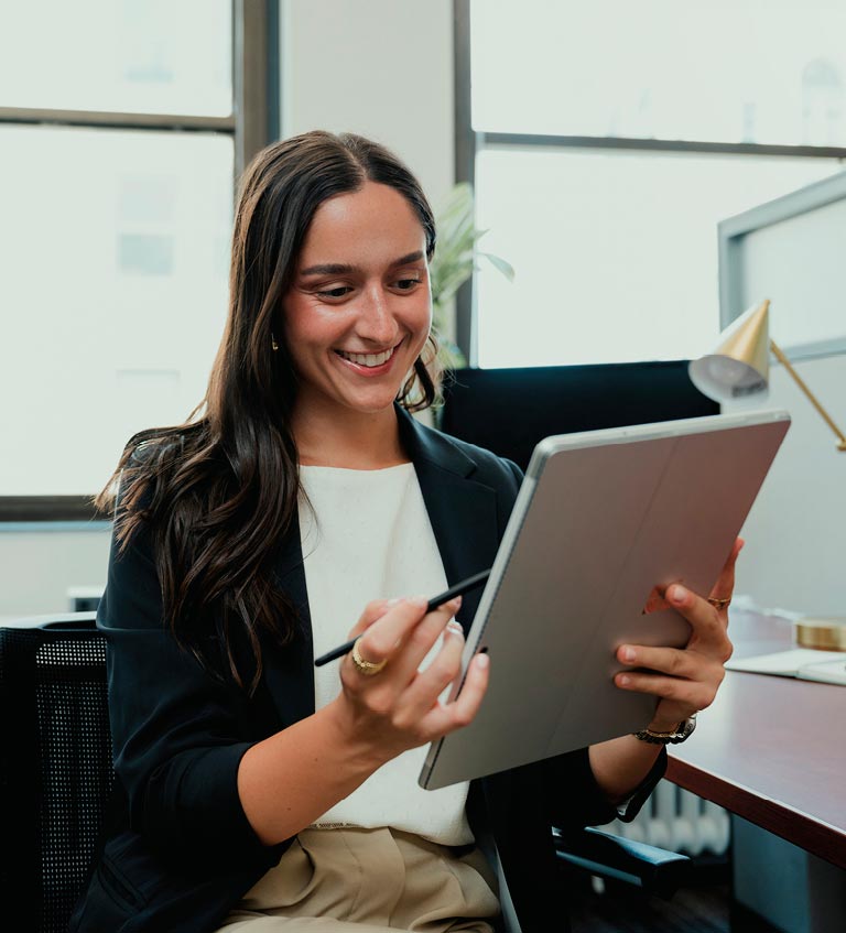 A woman sits at a desk in an office, smiling while using a stylus on a tablet.