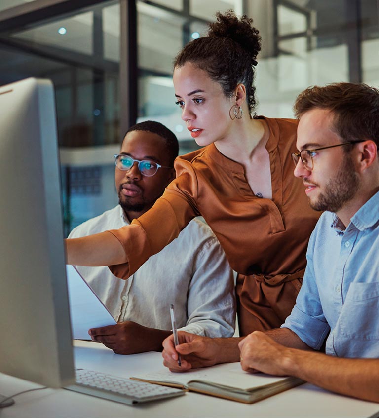 Three people in an office setting work together at a computer, with one person pointing at the screen and the others observing and taking notes.