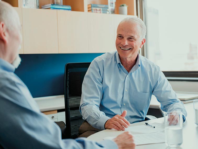 Two older men in dress shirts sit at a table in an office, having a conversation. One man is smiling and gesturing with his hand. Papers and a glass of water are on the table.