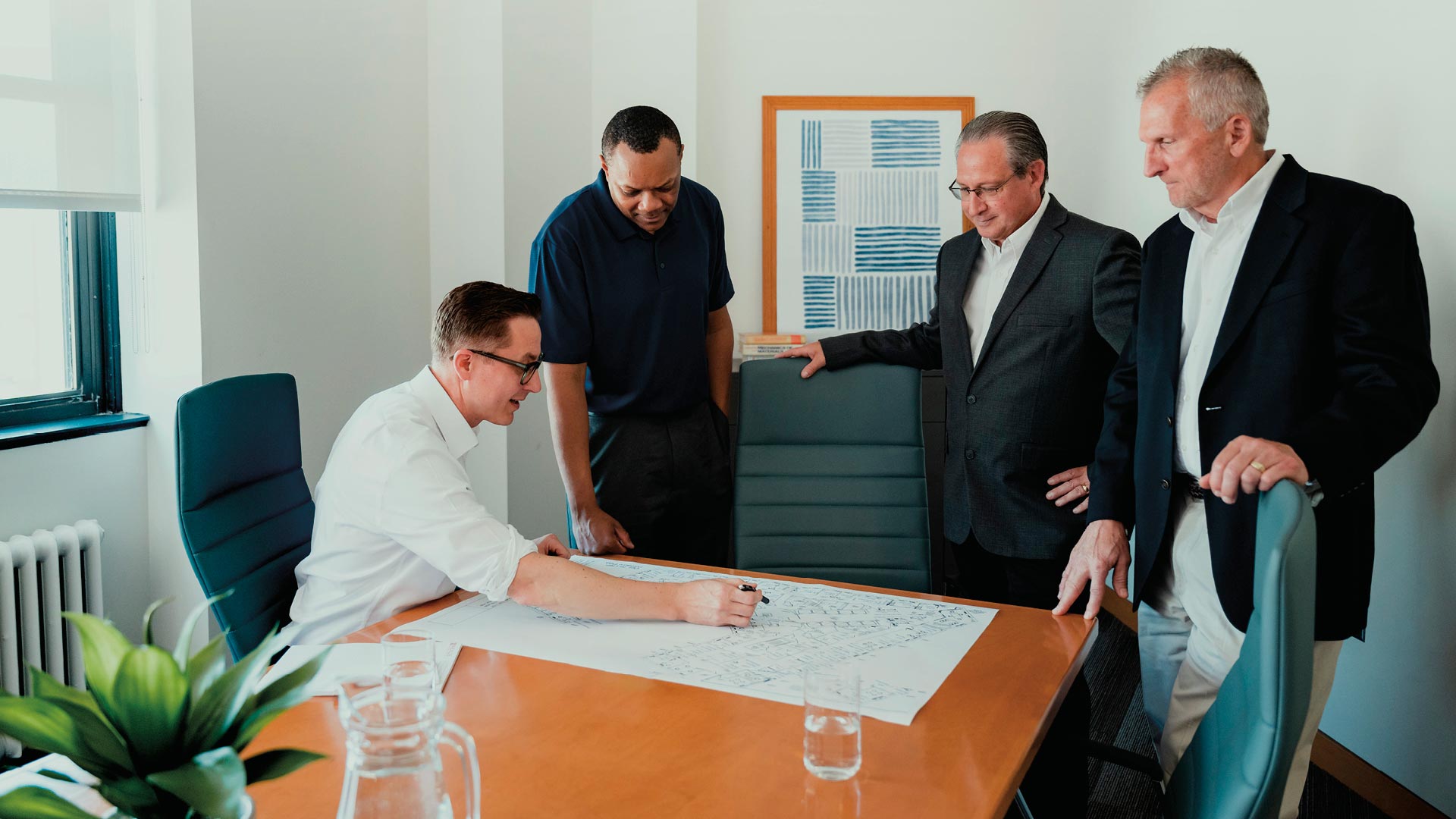Four men in business attire stand and sit around a conference table, reviewing and discussing documents and plans laid out on the table.