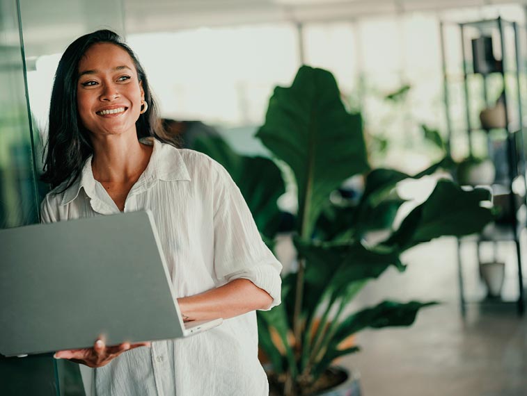 Woman in a white shirt holding a laptop, smiling and standing indoors near large green plants.