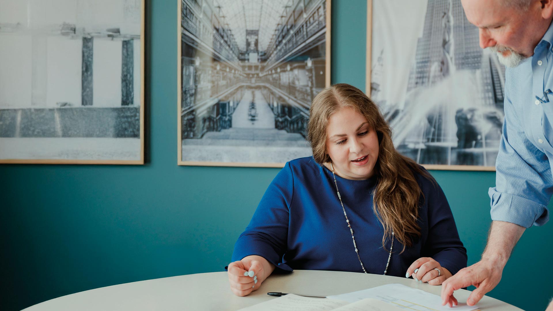 A woman sits at a round table with a notebook as a man stands beside her, pointing at the page. Framed architectural photos hang on the teal wall behind them.
