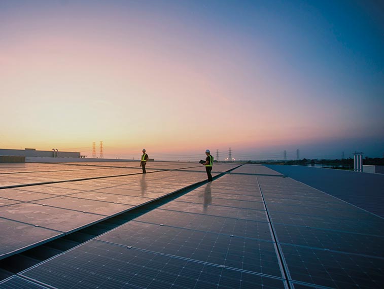 Two workers in safety gear stand on a large rooftop covered with solar panels at sunset, embodying the energy infrastructure of tomorrow, with power lines visible in the distance.