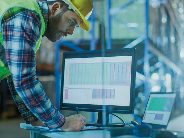 Man in safety vest and hard hat reviews data on a computer monitor in a warehouse, leaning on a desk with charts displayed on the screens, analyzing supply chain industry challenges.