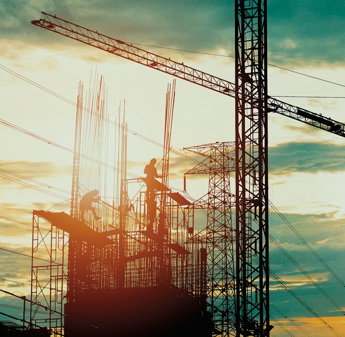 Silhouetted construction workers on scaffolding, surrounded by cranes and metal rods, work during sunset.