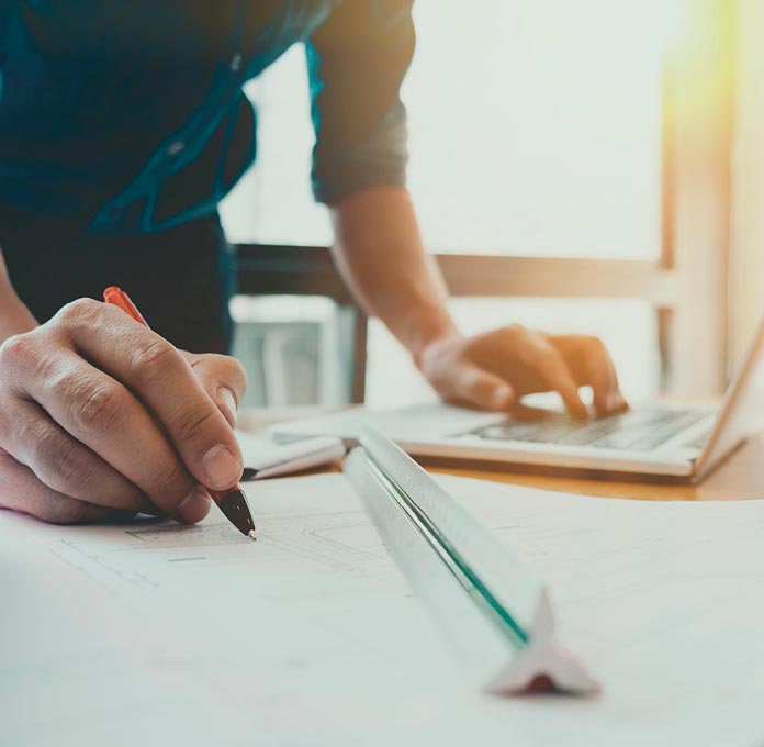 Person holding a pen, drawing on architectural plans with a ruler, while using a laptop in a sunlit office.