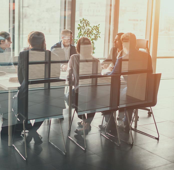 A group of people sit around a conference table in a glass-walled office, engaged in a meeting with sunlight streaming through the windows.