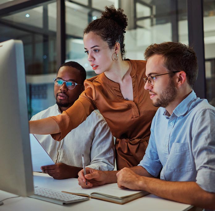 Three people work together at a desk, looking at a computer monitor. One person points at the screen while the others take notes.