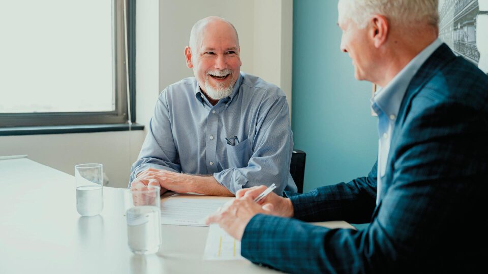 Two men sit at a table in an office, conversing and smiling. Both have documents and glasses of water in front of them. Natural light comes in from a window.