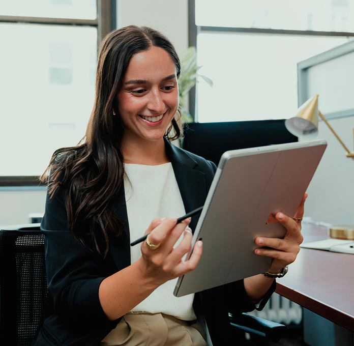 A woman sits at a desk in an office, smiling while using a stylus to write or draw on a tablet.