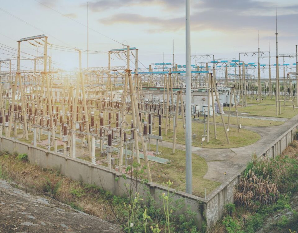 Close-up view of an electrical substation with rows of transformers, insulators, and metal frameworks connected by overhead power lines. The facility is enclosed by a low concrete wall, with grassy areas and service paths visible throughout. Tall utility structures and poles extend into the distance under a lightly overcast sky.