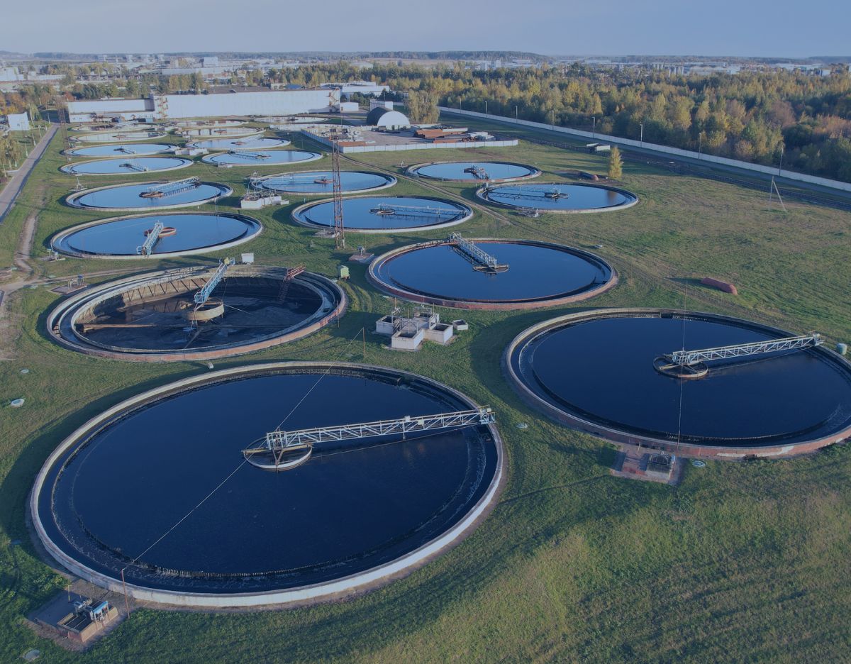 Aerial view of a wastewater treatment facility featuring multiple large circular clarifier and settling tanks arranged across a landscaped site. The tanks contain water at various treatment stages and are connected by access bridges, piping, and processing equipment. Industrial buildings and wooded areas are visible in the background.