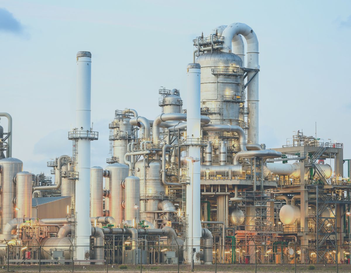 Industrial chemical processing facility with multiple distillation columns, stainless steel vessels, interconnected piping, and tall exhaust stacks. The large-scale plant is illuminated by soft daylight, highlighting the complex network of equipment and structures