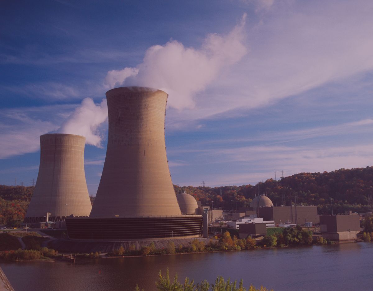 Large nuclear power plant beside a river, featuring two cooling towers releasing white steam into the sky. Reactor buildings and support structures sit along the waterfront, with wooded hills in the background under a partly cloudy blue sky.