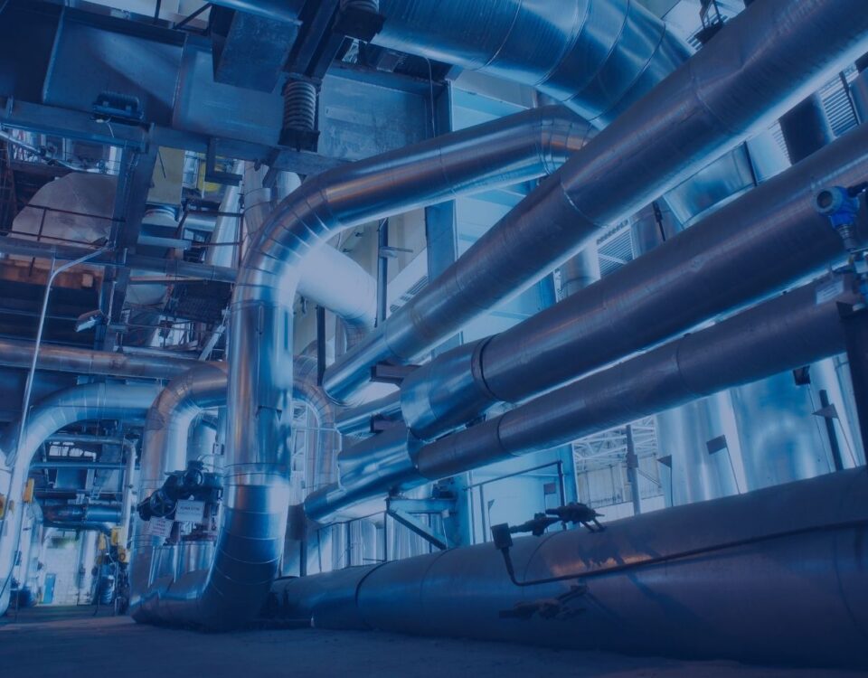 Large industrial facility with an extensive network of insulated metal piping and ductwork running overhead and along the floor. The image shows a complex mechanical system inside a power plant or refinery, with steel support structures, valves, and equipment visible throughout the brightly lit interior.