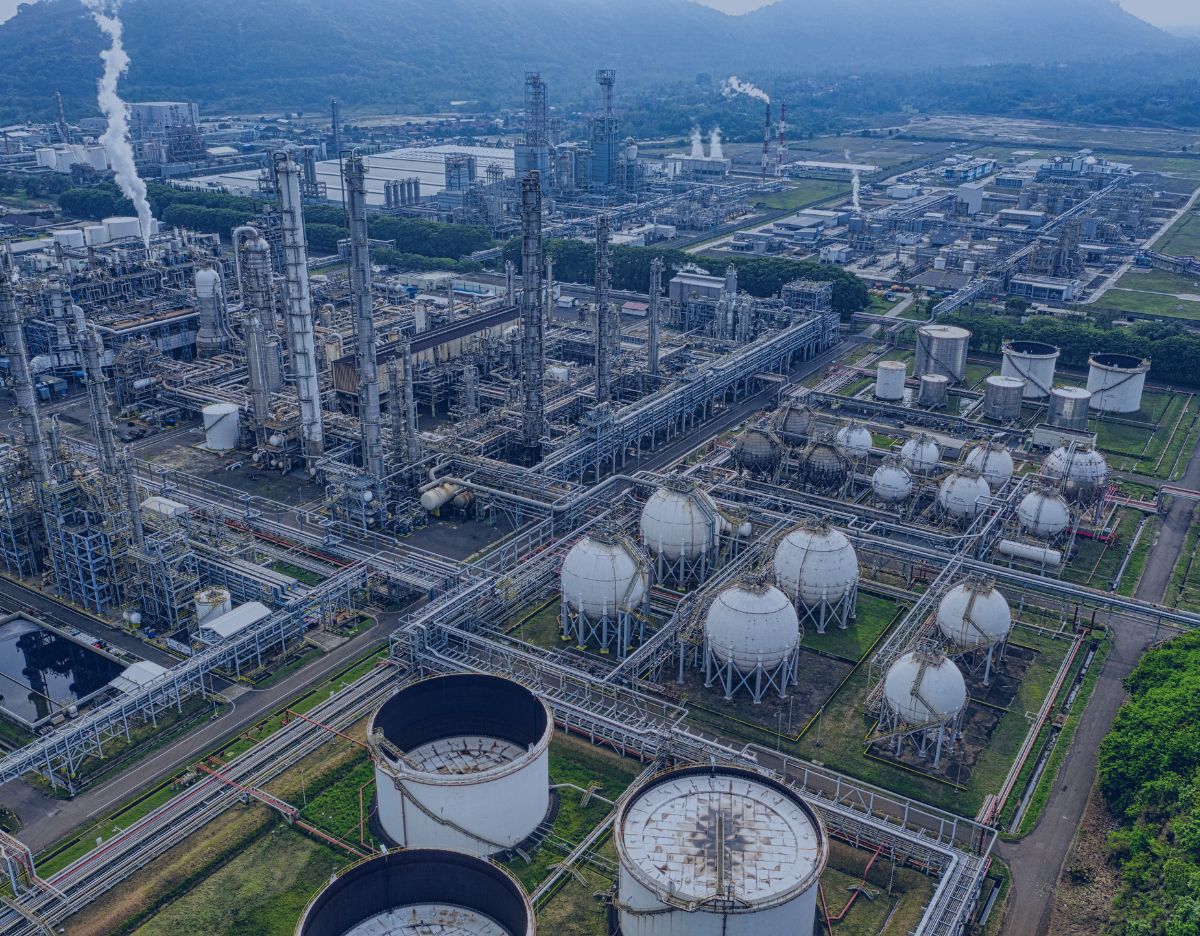 Aerial view of a large industrial processing facility with extensive piping networks, distillation towers, storage tanks, and spherical gas storage vessels. Steam rises from several processing units, while roads and utility infrastructure connect different sections of the complex. Mountains and additional industrial buildings are visible in the background.