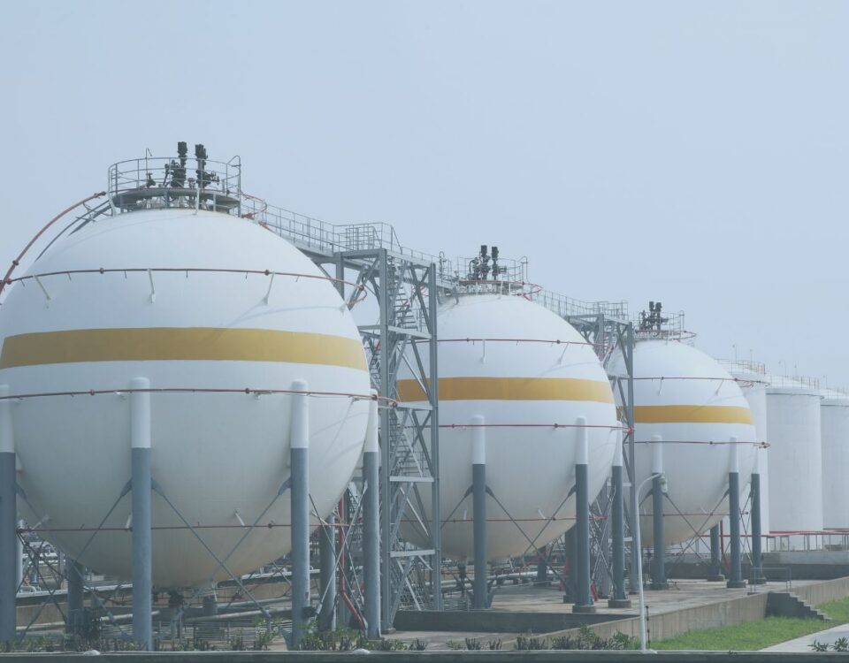 A row of large white spherical storage tanks with gold bands stands at a LNG facility. The tanks are elevated on support columns and connected by metal stairways, platforms, and piping. Additional cylindrical storage tanks are visible in the background under a hazy gray sky.
